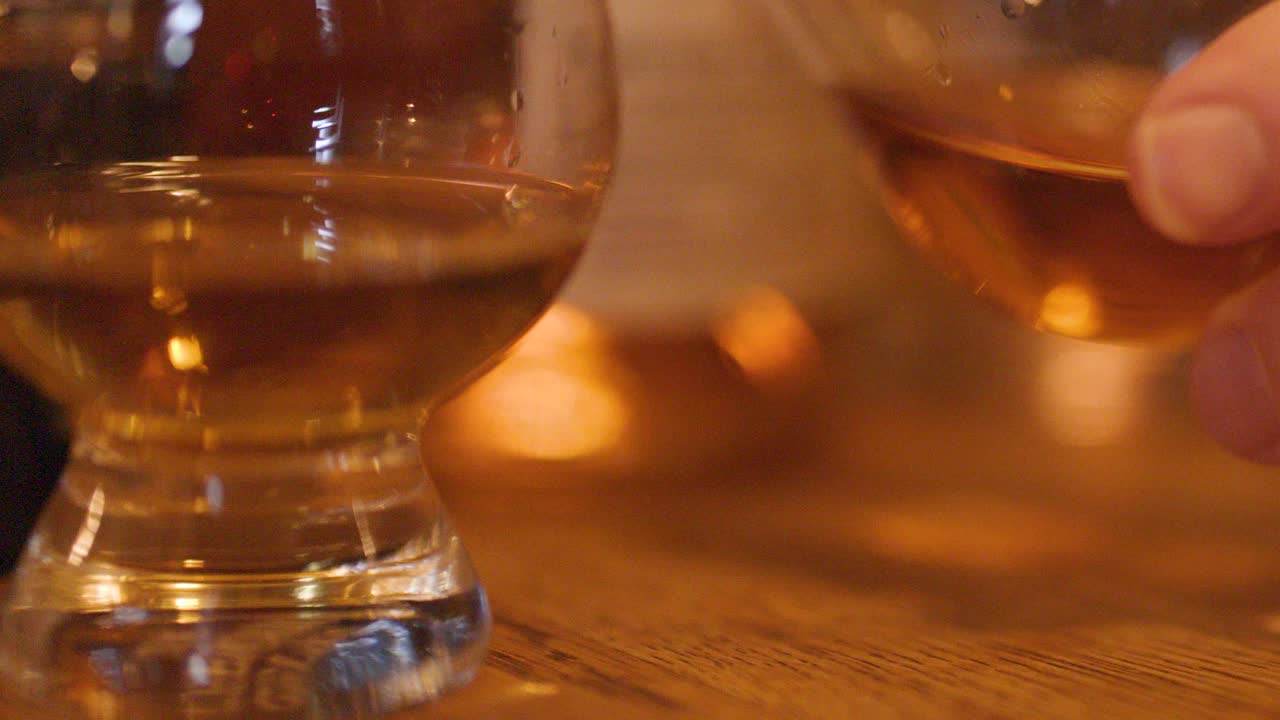 A hand reaches for a whisky glass on a wooden bar table in a warmly lit pub, with blurred bottles and ambient lighting in the background