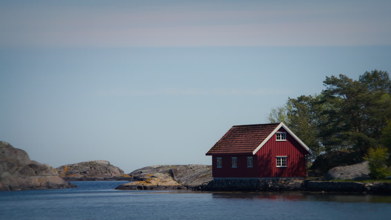 Nærbilde av rød hytte i tre ved vannet i Raet nasjonalpark i Norge, omgitt av trær og steiner