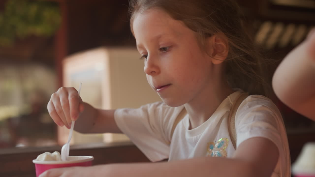 Concentrated little girl picks soft ice cream with plastic spoon from cup sitting in cafe. Junior schoolgirl eats healthy sundae for dinner in canteen