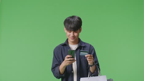 An Asian Shopping Man With Shopping Bags And Credit Card Using Mobile Phone Spending Money While Standing In Front Of Green Screen