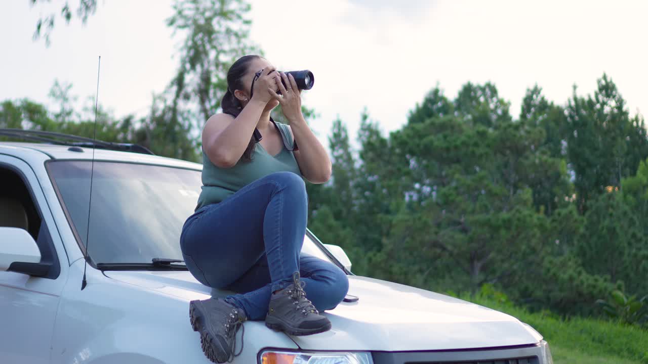 mujer fotógrafa de viajes, sentada en el capó de su coche todo terreno 4x4, con una cámara en sus manos, tomando fotografías al aire libre durante la puesta de sol