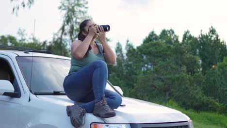 travel photographer woman, sitting on the hood of her 4x4 suv offroad car, with a camera in her hands, taking photographs outdoors during the sunset