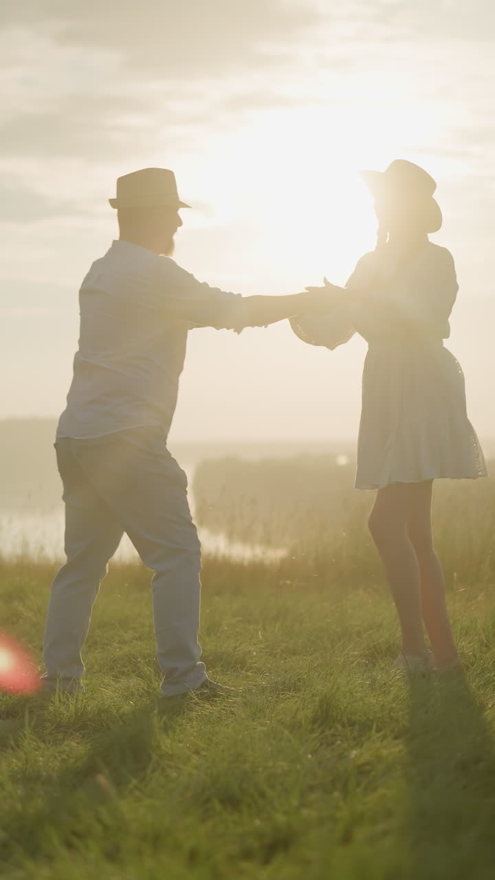 사랑하는 부부는 평화로운 호수 에 빛이 빛나는 잔디 언덕에서 춤을 춘다. 색 셔츠, 모자, 청바지를 입은 남자는 색 드레스와 검은색 모자를 입은 아내와 함께