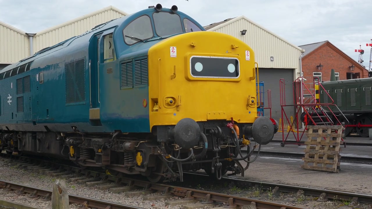 Et klassisk blåt og gult British Rail diesellokomotiv står stille på spor uden for et vedligeholdelsesskur i Gloucester, der viser kulturarv inden for jernbanetransport