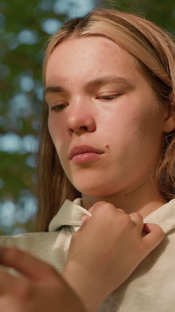 mujer joven con ropa casual opera atentamente el teléfono mientras sostiene su collar de capucha, la cara iluminada por la luz del sol filtrando a través de las hojas, el fondo borroso con vegetación