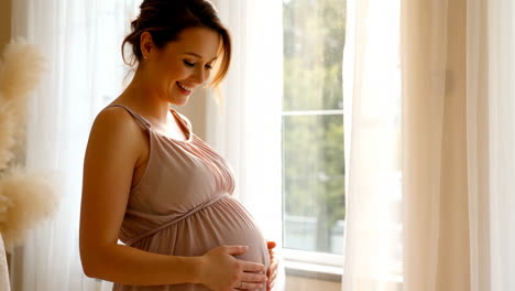 Happy pregnant woman gently touching her baby bump by a window