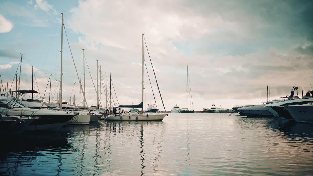 A sailboat approaches a marina surrounded by yachts and calm, reflective water under a cloudy sky
