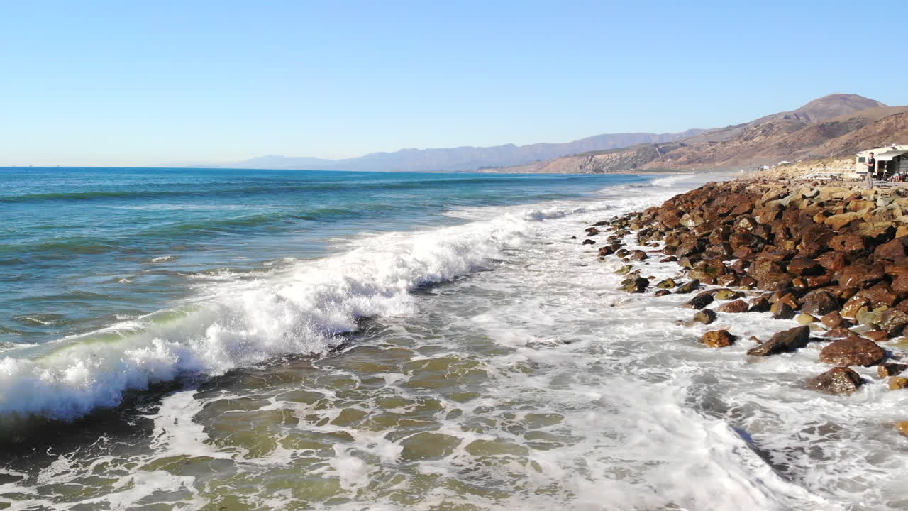 Epica ripresa aerea con drone da un'onda oceanica blu che si infrange a una vista che domina le spiagge dorate della costa californiana e le palme.