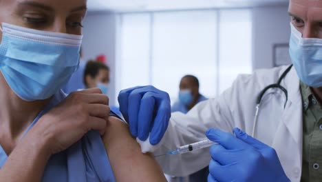 caucasian male doctor giving covid vaccination to female colleague at hospital, both in face masks