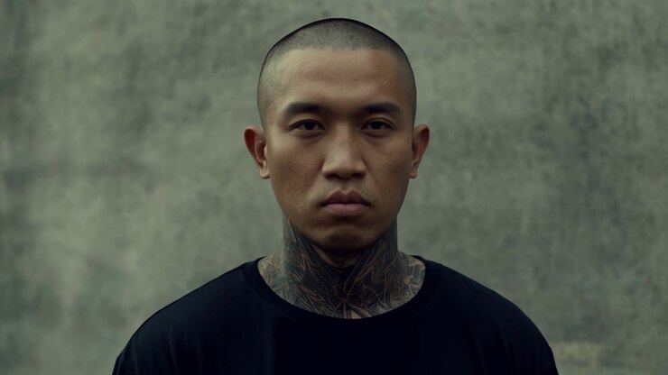 Close Portrait Asian Man With Tattoos, Shaved Head And Neck Ink Against Weathered Concrete Wall, Neutral Stoic