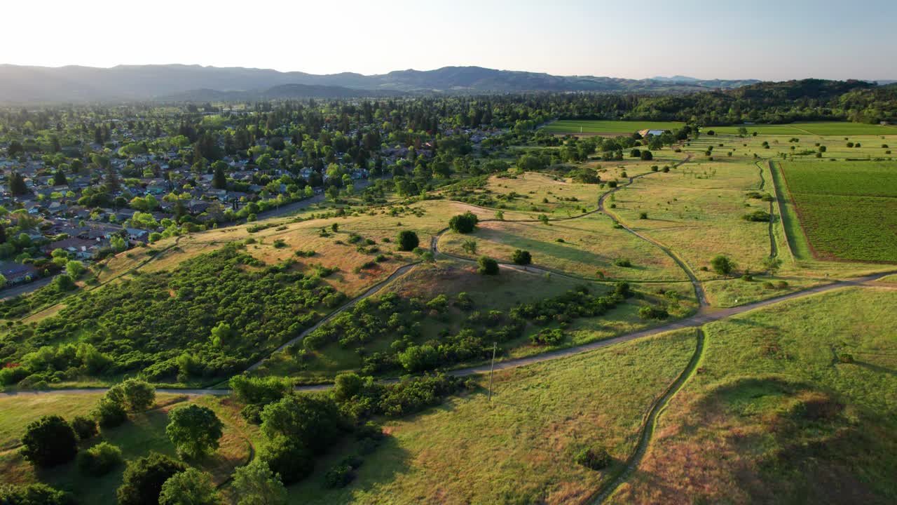 Napa Valley, California. Slow aerial panoramic pan over lush, vibrant green rolling hills along a hiking trail near vineyards, as the sun casts beams over the town below.