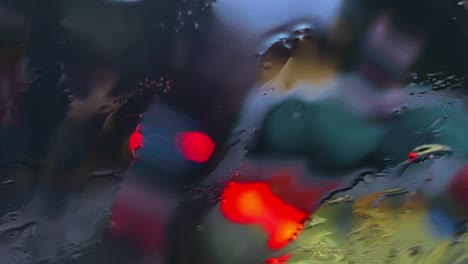 Close-up-of-raindrops-and-sleet-sliding-down-on-tarnished-windscreen-and-blurred-lights-of-car-city-traffic-in-background