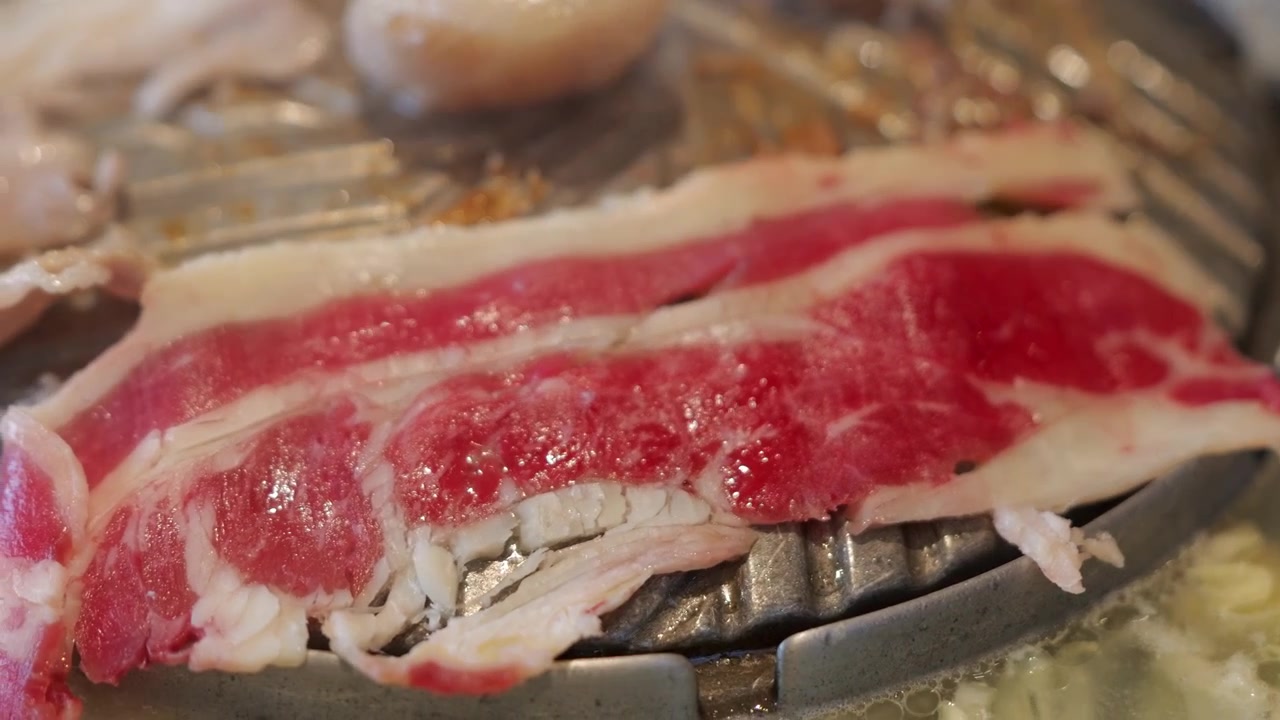 熱いタイ式バーベキュー鍋で焼かれる薄切り牛肉のクローズアップ。ジュージューと音を立てる肉、立ち上る煙、赤々と燃える熱、食欲をそそる焼き目を捉える。