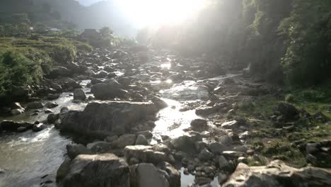 stunning aerial flyover of a rocky river during sunset and golden hour with crystal reflections of the sunlight in the river water and surrounded by green mountain landscape