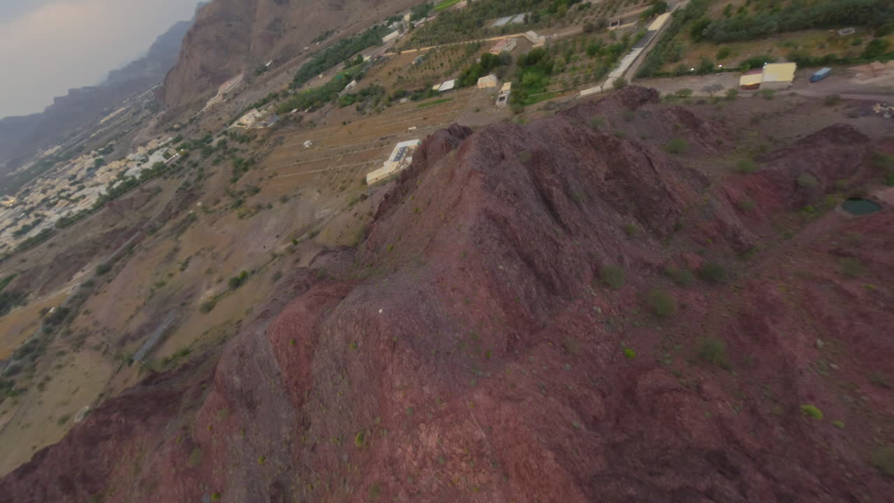 FPV drone turns above rocky hill then speeds over palm groves and village houses at desert edge