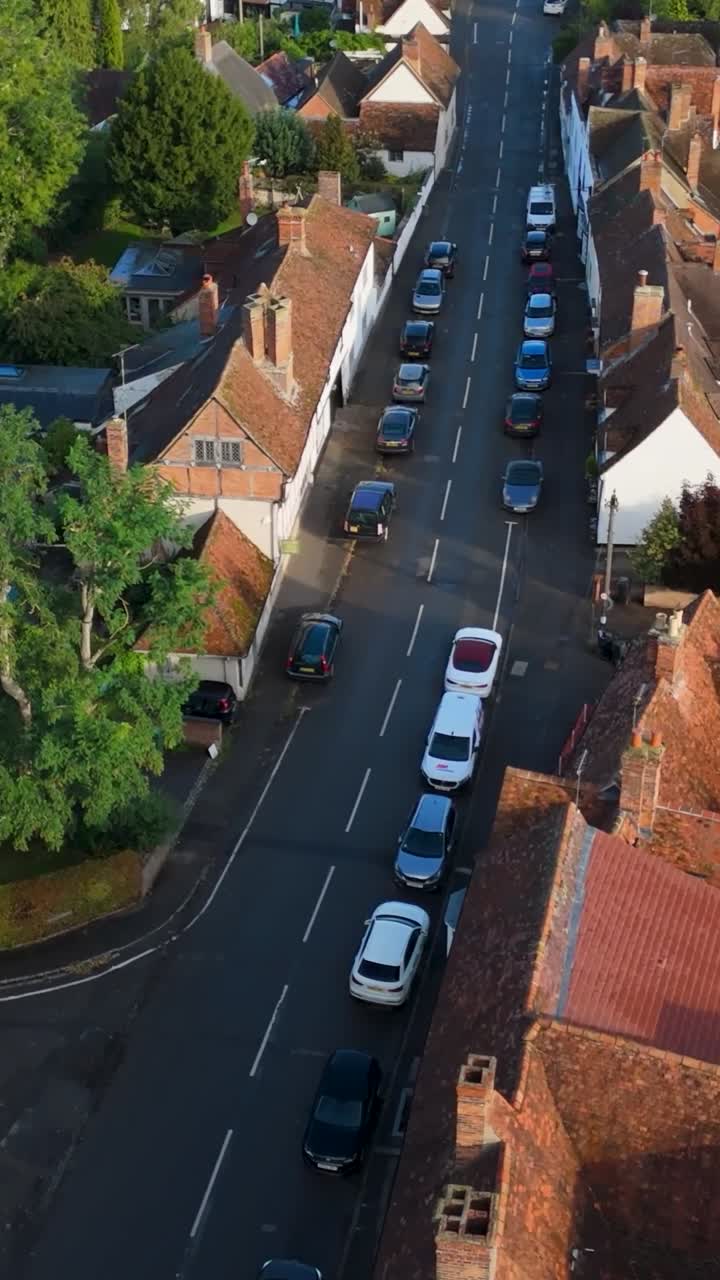 Vertical drone flight over Dorchester on Thames village. Follows the curved road, showing charming old rooftops, cars, and leafy trees framing this picturesque Oxfordshire English countryside scene