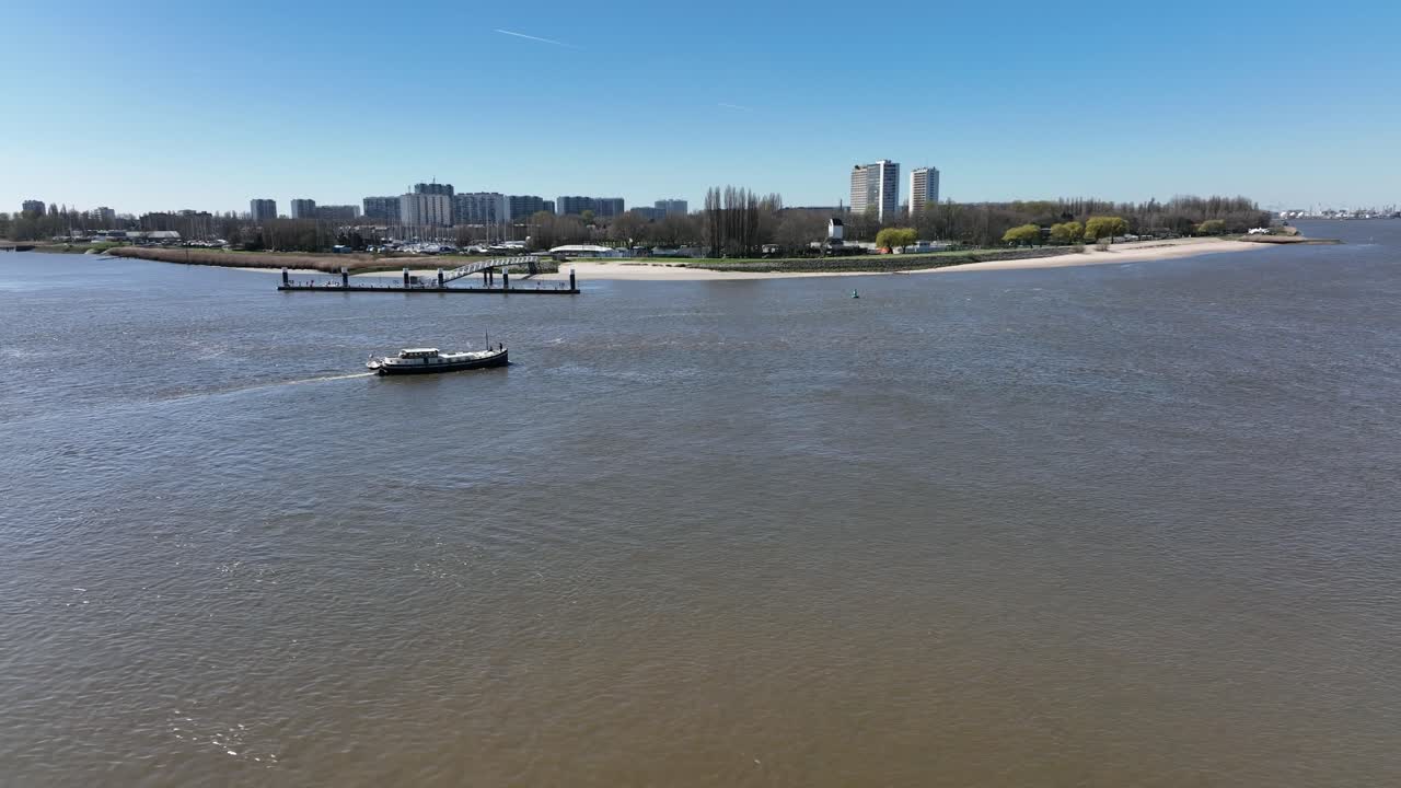 Semi orbit aerial shot of boats cruising Antwerp river with residential towers, green riverside, and cityscape skyline in background