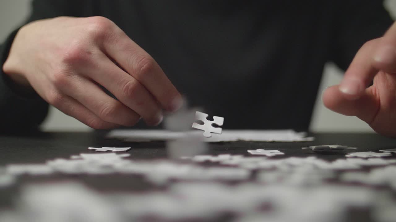 Person's hands trying to solve a puzzle in the background with white puzzle pices falling on a dark table simultaneously. Shot in 4K, Slow Motion with Shallow Depth of Field