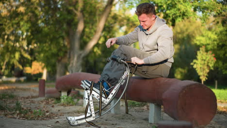 young man sitting on reddish-brown log in park wearing light gray sweatshirt and khaki pants, focused on attaching straps to spring stilts beside him, surrounded by green trees