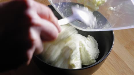un primer plano de un tazón de mezcla negro sobre una mesa de cocina de madera, un chef que prepara cuidadosamente queso blanco bajo en grasa transfiriéndolo de un recipiente de almacenamiento y colocándolo en el tazón