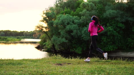 brunette with long hair in headphones runs along the river in the park in the morning at sunrise in the summer in a pink jacket and black pants