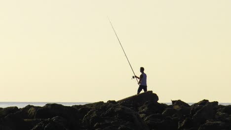 joven lanzando caña de pescar desde las rocas al amanecer, silueta de cámara lenta