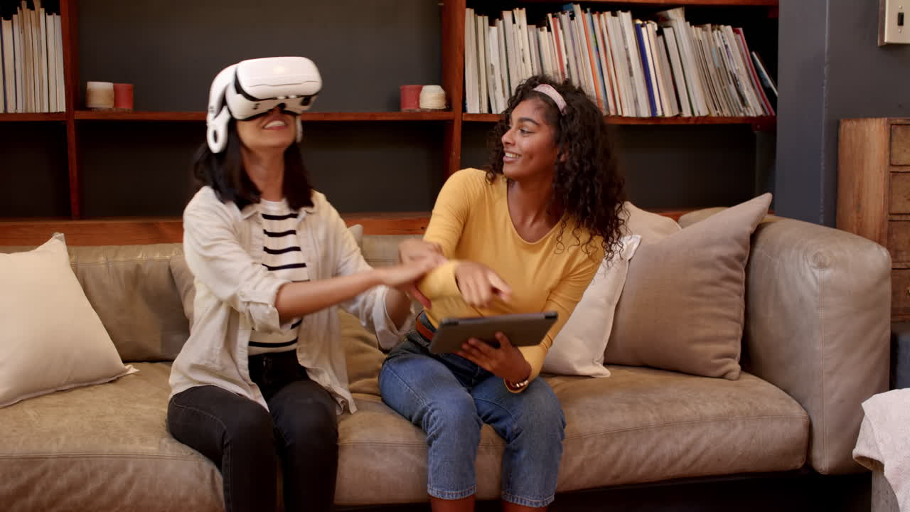 Multiracial female friends enjoying virtual reality and tablet on cozy living room couch, at home