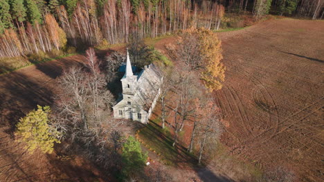 Drone-arching-shot,-of-a-white-little-countryside-church-surrounded-by-fields-and-forest-in-autumn