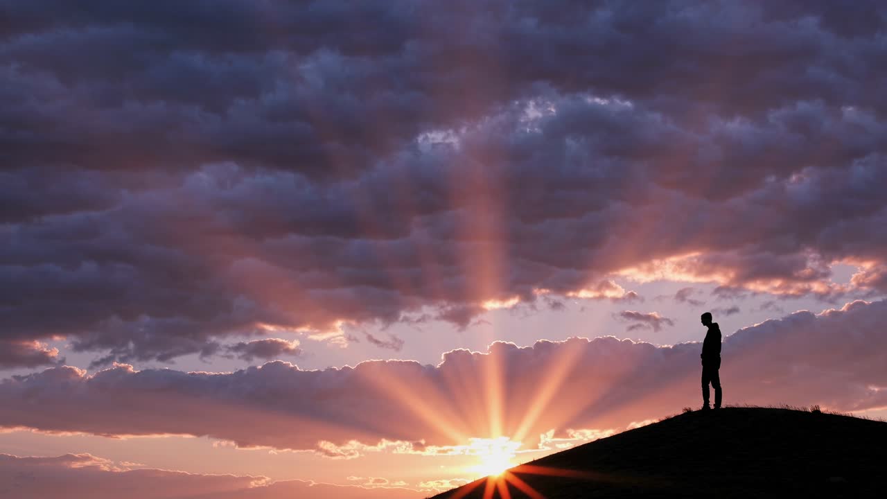 Silhouette of a person on a hill at sunset, with dramatic sun rays breaking through clouds