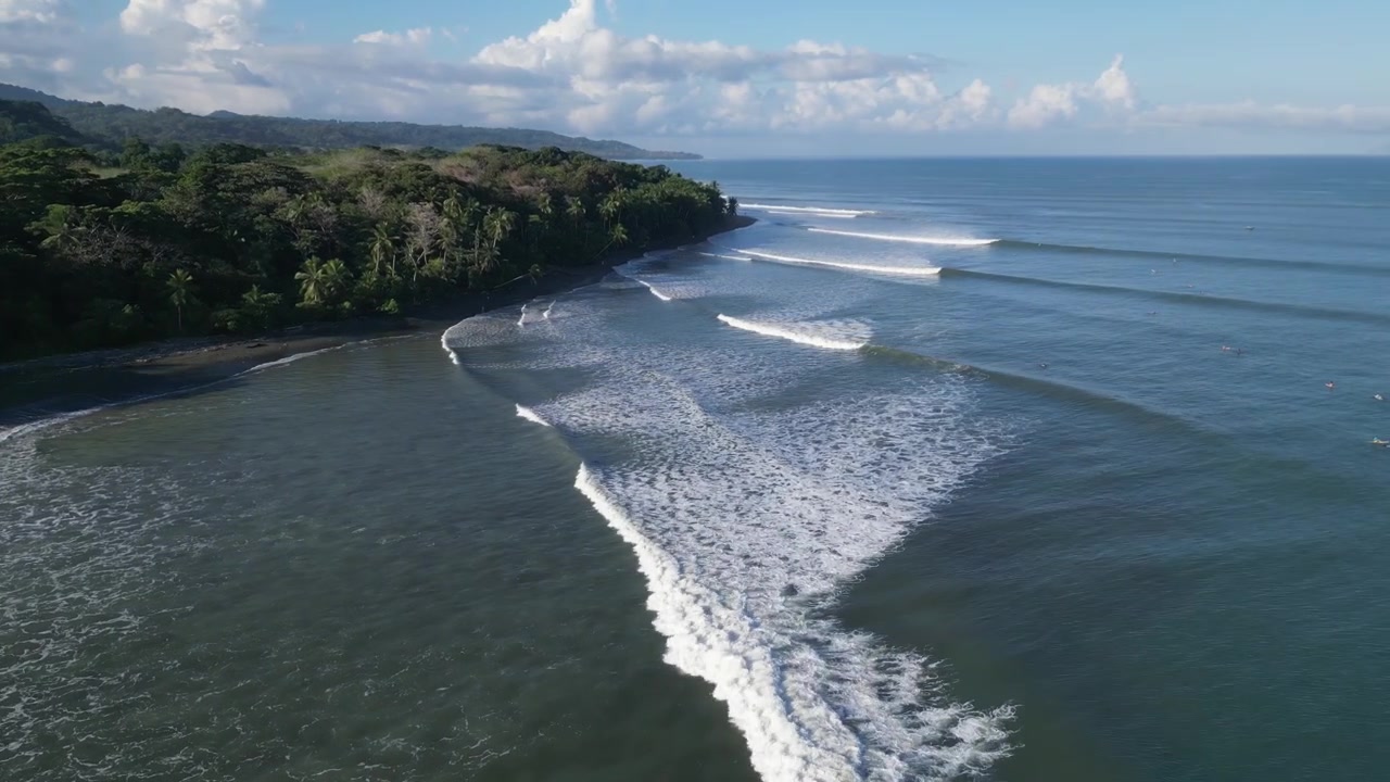 Ripresa cinematografica con drone che scende più vicino alla superficie dell'oceano, angolata verso la costa, tracciando una linea d'onda incredibilmente lunga e perfetta che si srotola lungo una lussureggiante spiaggia tropicale in Costa Rica.