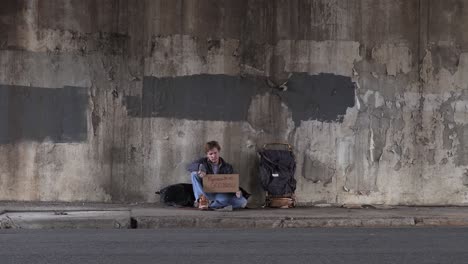 Wide-Shot-of-a-Homeless-Man-Sitting-on-the-Sidewalk-Holding-a-God-Bless-Cardboard-Sign