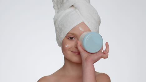 Young-smiling-child-girl-applying-cleansing-moisturizing-cream,-looking-at-camera-holding-creme-jar