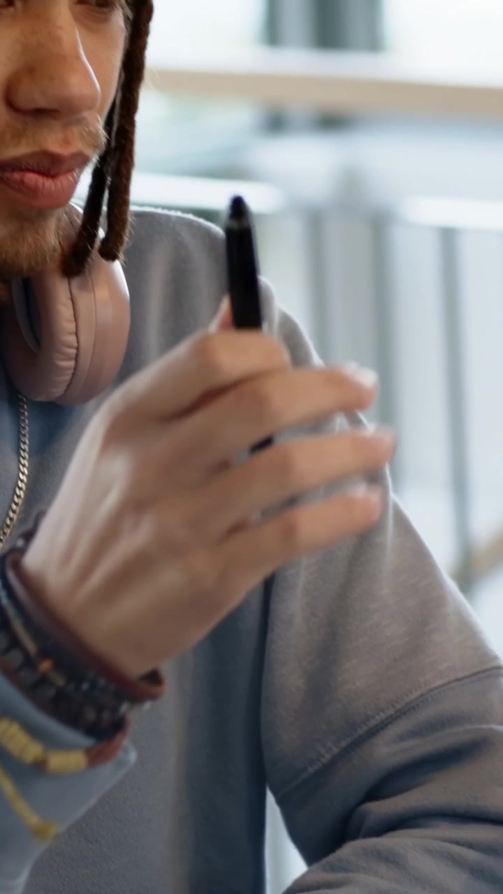 Vertical video: Glancing down, man in blue sweater tapping pen, studying in cafe with pink headset