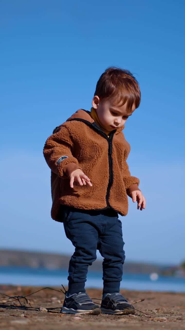 Cute kid walks by the waterfront looking for the stones. Baby boy touches a twig and then continues to go. Low angle view. Vertical video