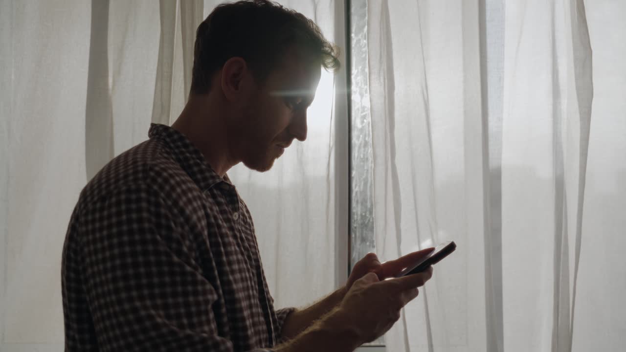 Texting on smart phone. Man standing indoors by window with white curtains, focused on phone. Soft daylight, calm and intimate. Technology, texting, browsing, social media, online communication