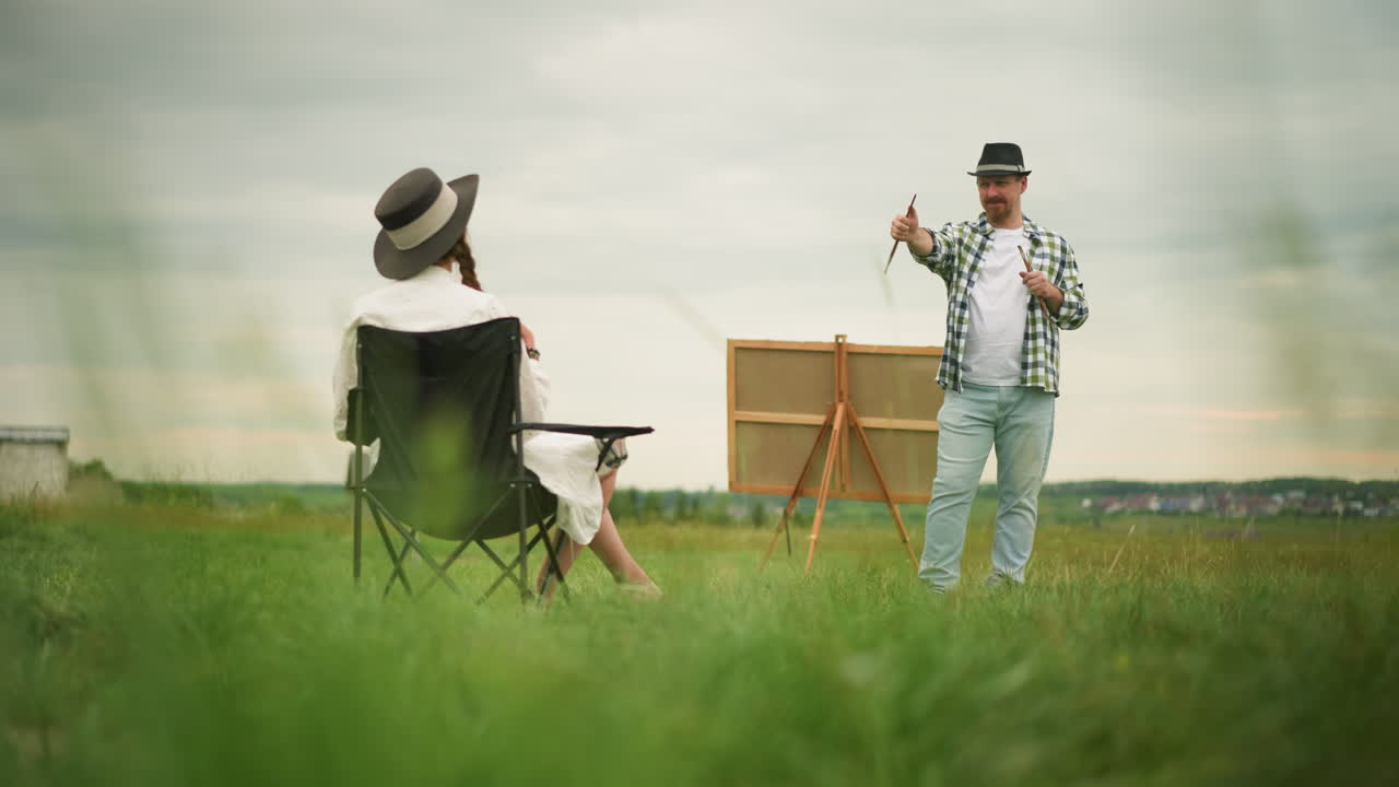 체크 된 셔츠와 청바지를 입은 조각가는  옷과 모자를 입은 여자를 향해 브러쉬를 가리키며 미소 짓고, 잔디에 있는 의자에 앉아 있습니다. 큰 보드는 배경에 설정되어 있습니다.