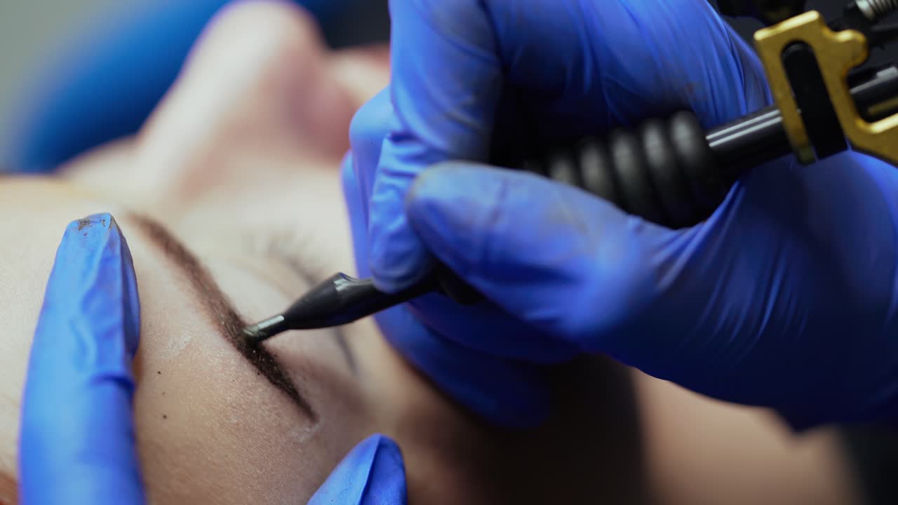 Beauty master makes eyebrow tattoo in blue gloves with the use of a special needle and a pistol in the salon. Close-up