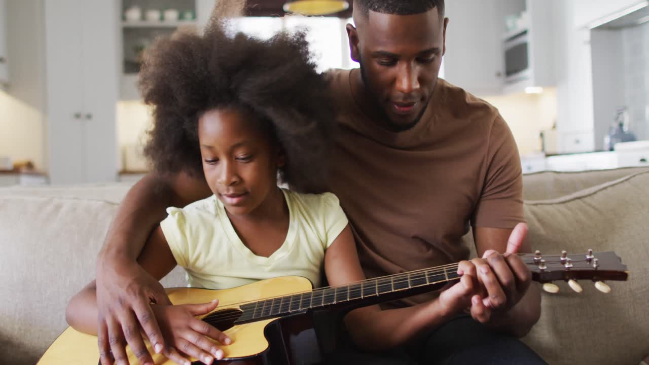 Padre afroamericano e sua figlia seduti sul divano che suonano la chitarra insieme.