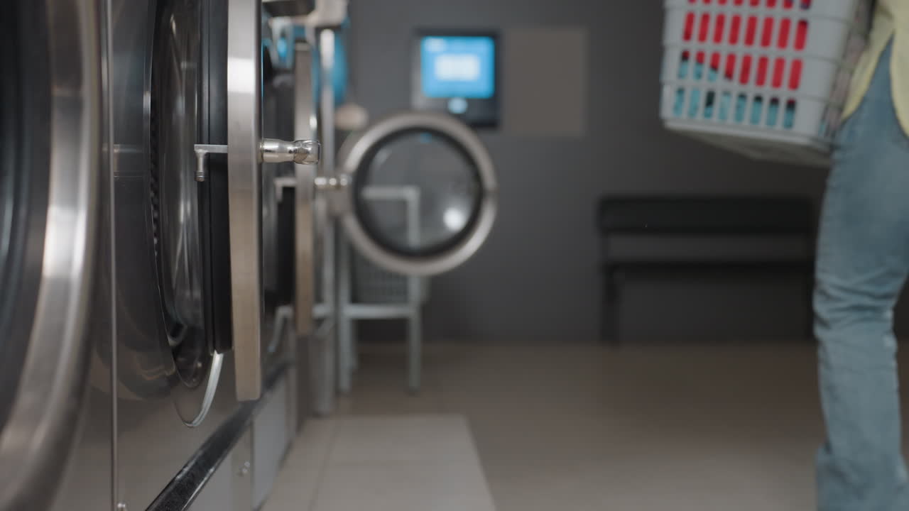 Vista inferior de una trabajadora que lleva una cesta de ropa mientras sale de una lavadora industrial dentro de una lavandería, puerta abierta, toallas a la vista, fila de máquinas, servicio de rutina, instalación limpia, funcionamiento sin problemas