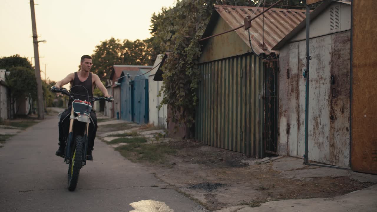 회색 티셔츠에 짧은 헤어을 가진 남성 오토바이 운전자는 자전거를 타고 다양한 도구로 가득 찬 그의 차고 작업장에 들어갑니다.