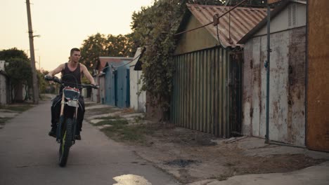 A-male-motorcyclist-with-a-short-haircut-in-a-gray-T-shirt-rides-his-bike-into-his-garage-workshop-which-is-filled-with-various-tools-and-he-puts-his-bike-on-a-jack