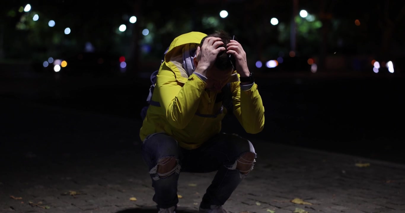 Turista smarrito con la giacca si accovaccia con espressione triste di notte.