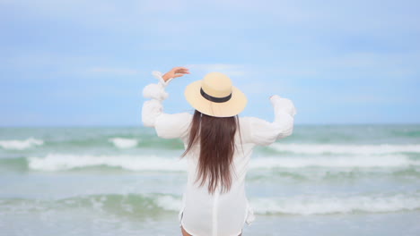 Frau-In-Weißer-Bluse-Steht-Am-Strand-Am-Meer-Und-Hebt-Die-Arme-In-Richtung-Endloser-Meereslandschaft-–-Urlaubsvorlage-Von-Hinten