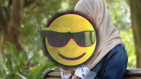 animation of happy emoji over woman in hijab using smartphone