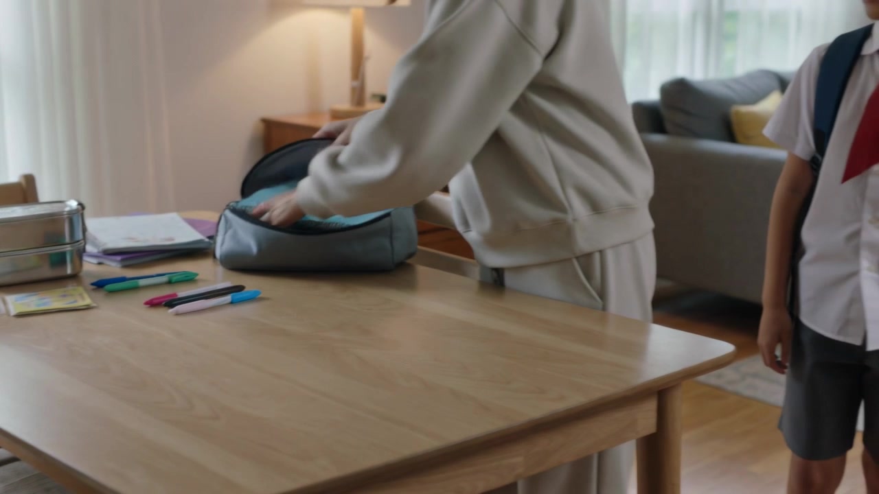 Packing parent in sweatshirt placing notebook into child's gray backpack for school at home table