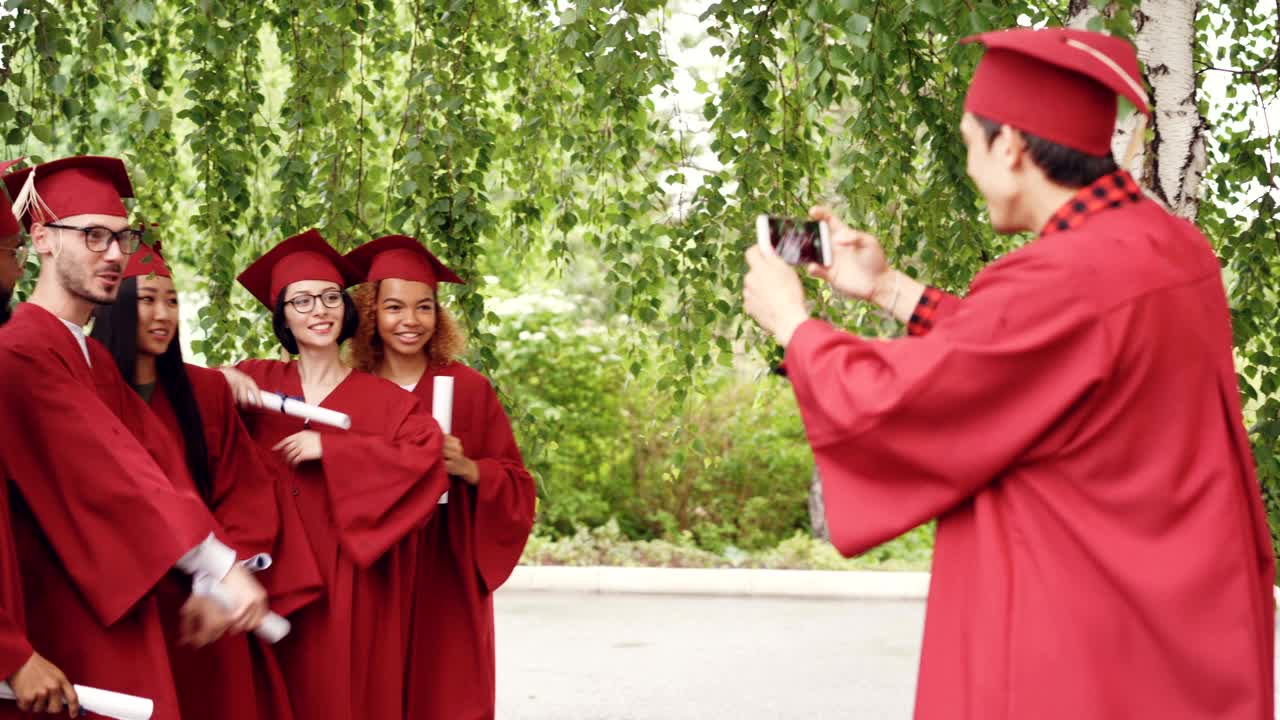 Giovane uomo con smartphone che scatta foto di laureati che si divertono posando con i diplomi, muovendo mani e dita e gridando di felicità. Concetto di istruzione e gioventù