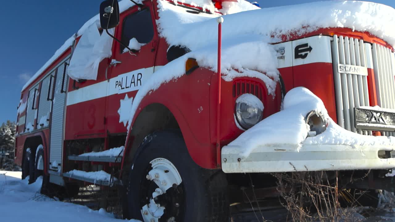 Low angle close up or closeup footage of a vintage retro red and white firetruck left abandoned in snow during a winter sunny day in a field that has white fluffy snow all around on the ground.