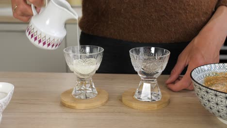 woman pours milk from vintage jug in crystal glasses, preparing dalgona coffee