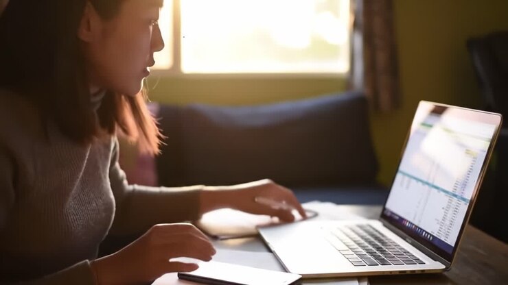 Fokuseret ung kvinde fordybet i arbejde på bærbar computer, tager noter med pen derhjemme, indfanger essensen af produktivitet og koncentration i et hyggeligt miljø