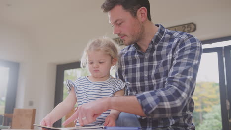 Padre-En-Casa-En-El-Mostrador-De-La-Cocina-Ayudando-A-Su-Hija-A-Leer-Un-Libro---Filmado-En-Cámara-Lenta